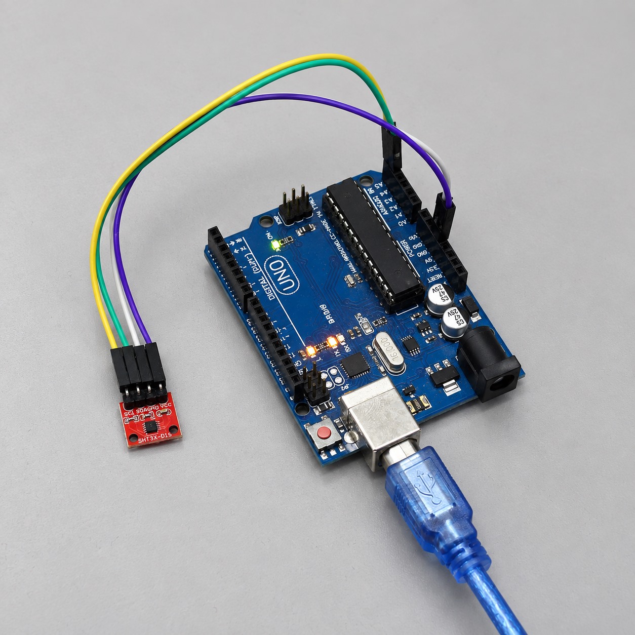 Arduino Uno connected to a red SHT31 breakout with four jumper wires and a USB cable on a clean electronics bench